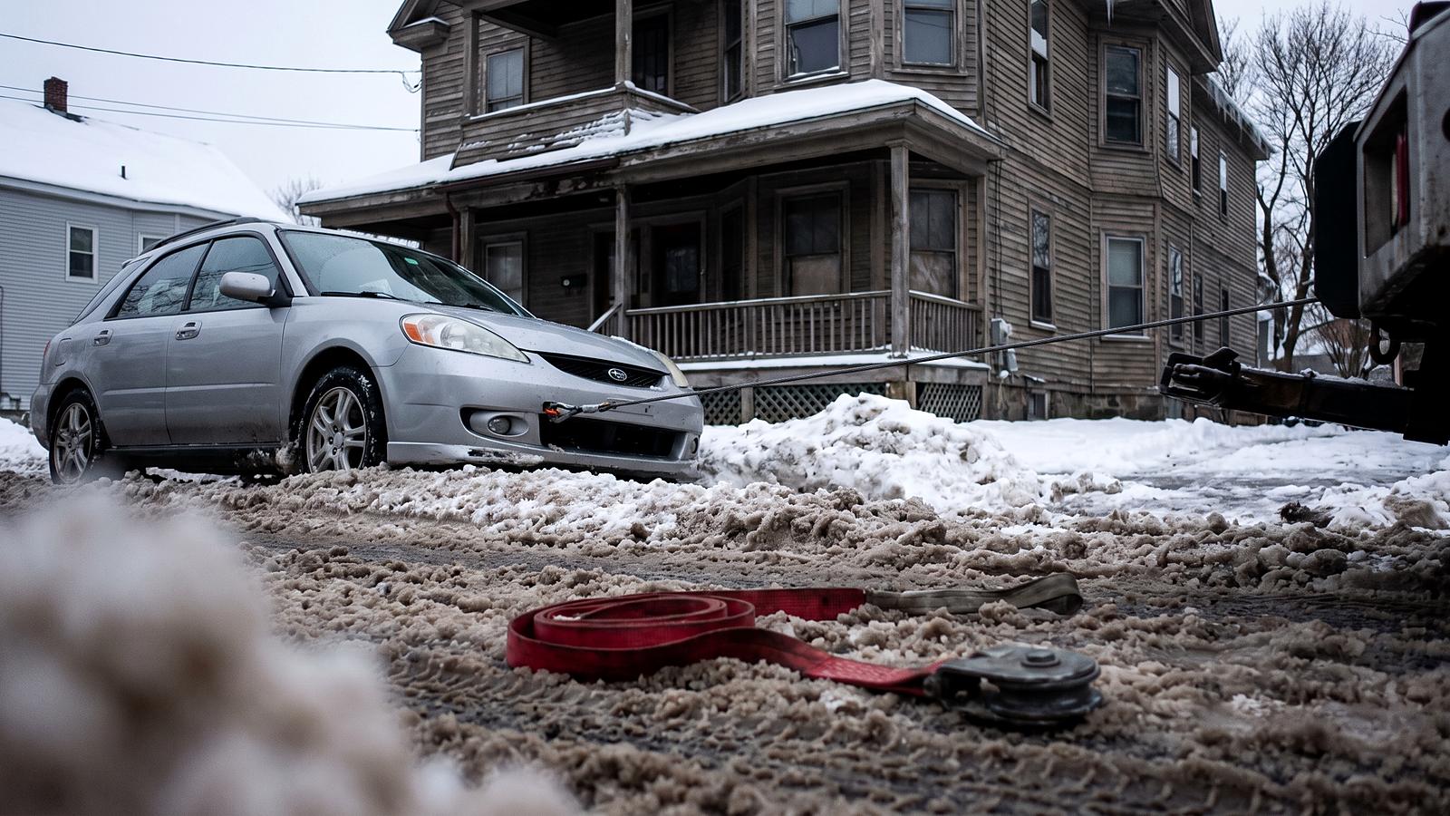 A silver car nosed into a plowed snowbank on a residential street, a red recovery strap and a snatch block lying in the slush